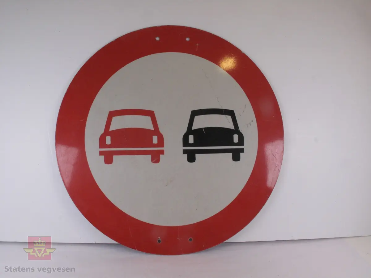 Sirkelformet skilt med rød bord (7,5 cm). Bunnen er hvit og lysreflekterende, og har figurer av en rød og en svart bil ved siden av hverandre. Den ene er rød, den andre svart. Skiltet betyr at forbikjøring er forbudt. Det har to skruehull på midten oppe og nede. Noen riper og litt fastgrodd skitt.