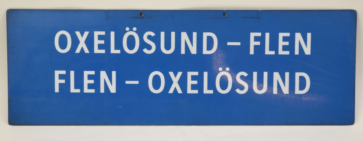 En blå avlång dubbelsidig destinationsskylt som har den vita texten "OXELÖSUND - FLEN FLEN - OXELÖSUND" på båda sidor.