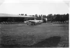 Ett amerikanskt bombflygplan - Consolidated B 24 Liberator - har landat på ett gräsfält.  I förgrunden vingspetsen av ett annat flygplan och bakgrunden vad som ser ut som stridsvagnshinder.