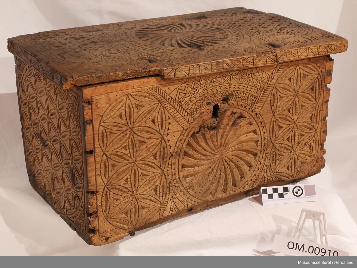 Mellomstor trekvit kiste i furu, rikt dekorert med skoren ornamentikk midt på loket, på bak- og frontsida, ellers rikeleg rosettornamentikk. Utskjeringane skal syne dei 4 årstidene. Skrinet luktar kamfer. Naglane fungerer som ei slags sikring. Skrinet har to jernhengsler og beslag på innsida av loket.