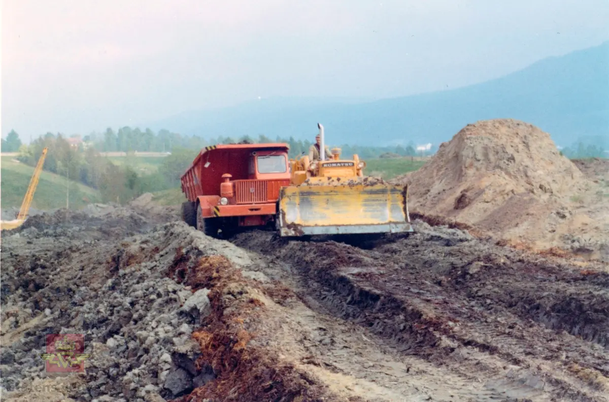 Veganlegg Vinnes - Krokstadelva i Nedre Eiker. Planeringsarbeid. Transport med dumper, planering med Komatsu bulldozer. "Bløt" byggegrunn. 
I bakgrunnen en rød tipptruck, Kockum Landsverk . (Informant: Jo-Olav Bakken)
Se Relasjon og gjenstandsnummer NVM 00-G-02329 for mer informasjon om anleggsmaskinen fra Kockum Landsverk.
Usikkert når bildet er tatt. 
Dette bildet må være tatt før 1975. Antatt 1965-1970. (Informant Brynjar Aasen)