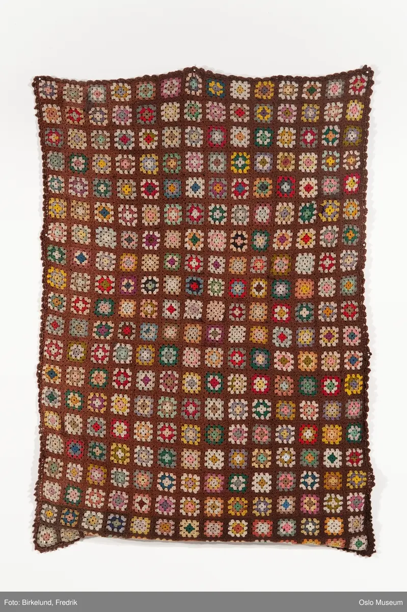 Heklet lappeteppe i brun farge med  forskjellige fargemotiv i de forskjellige lappene. Lappene er ca 7/7 cm. Teppet er trukket på den ene siden med  lys brunt stoff. Heklet kant rundt  teppet i mørk brun.