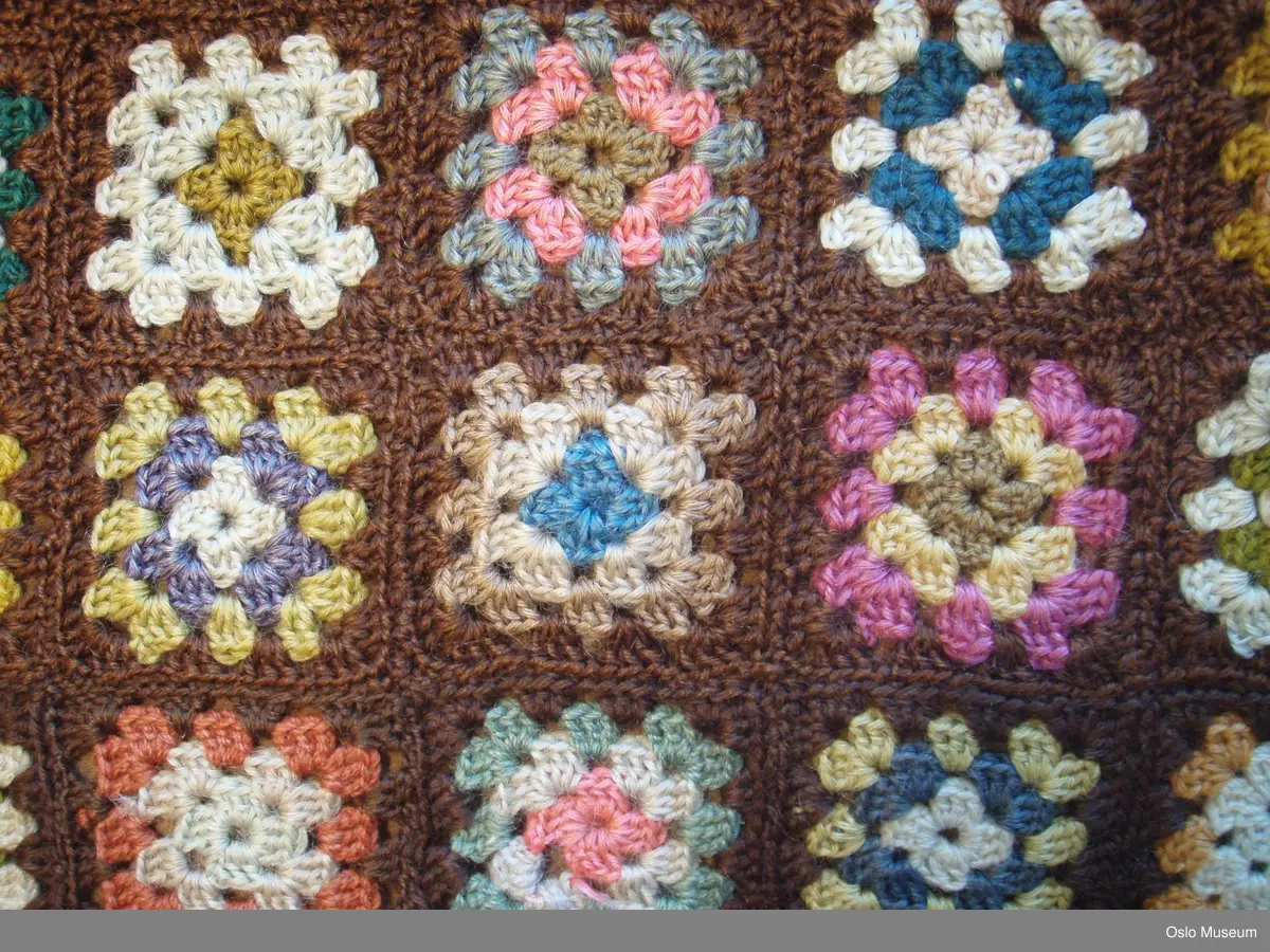 Heklet lappeteppe i brun farge med  forskjellige fargemotiv i de forskjellige lappene. Lappene er ca 7/7 cm. Teppet er trukket på den ene siden med  lys brunt stoff. Heklet kant rundt  teppet i mørk brun.