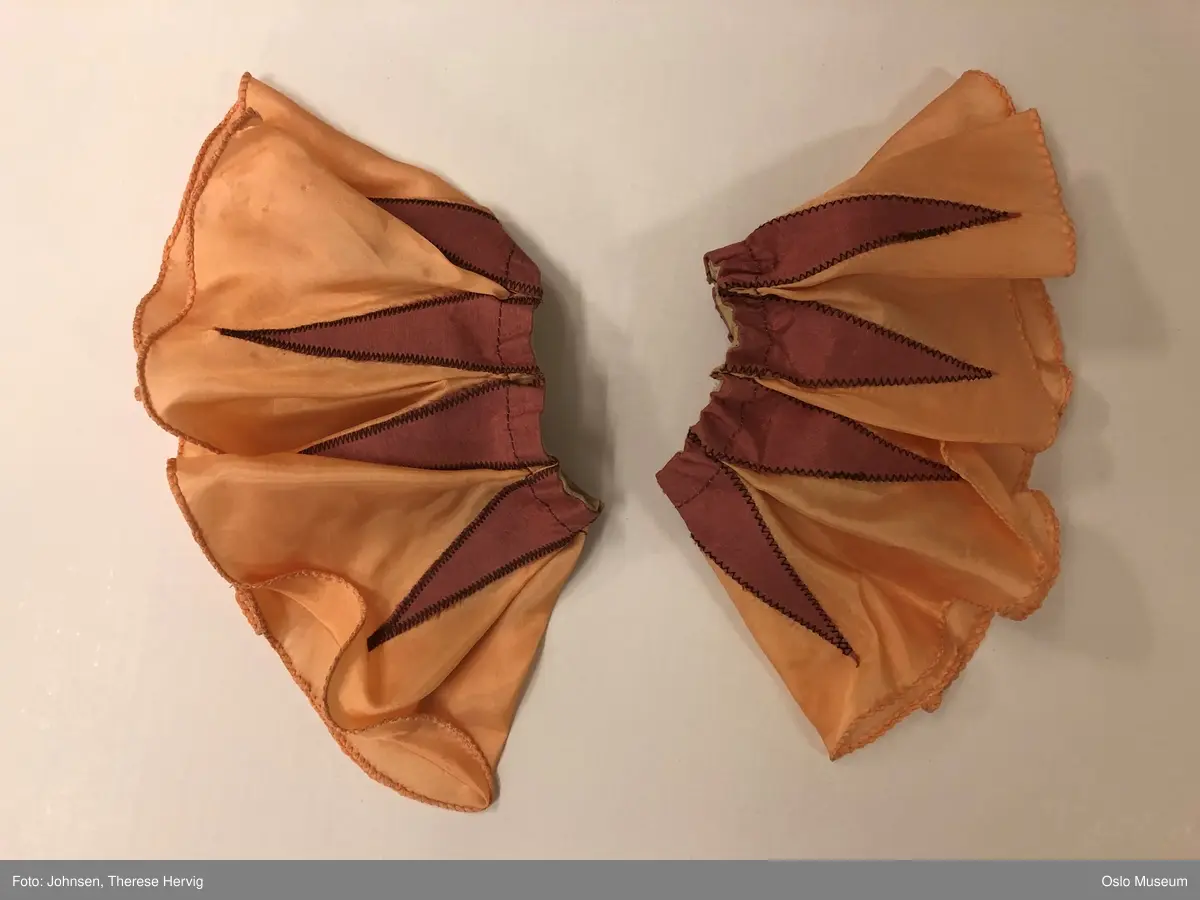 Todelt ballettkjole som holdes sammen med knapper.
A Innsvingt liv, holdes sammen med hekter i ryggen. Tynne skulderstropper i strikk. Blek rosa/ ferskenfarget bunn med innfellinger i mørkere rosa. Utringing er dekorert med grønn kant. 
B: Strutteskjørt i blek rosa, fersken med innfellinger i mørkere rosa. Dekorert med applikerte blomster i rosa, gul og brun. Mørk rosa bånd i livet som knytes i sløyfe på ryggen. Skjørtet lukkes med hekter og knapper i rygg. 
C: hodepynt, avstivet, kledd i samme tekstil som resten av kostymet. Dekorert med rosa paljetter. 
D: ferskenfarget erme med innfellinger i mørkere rosa. 
E: ferskenfarget erme med innfellinger i mørkere rosa.