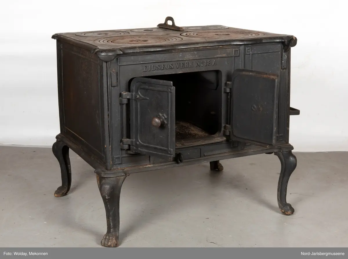 Stor, firkantet komfyr med dobbel ovnsdør til stekeovn? med støpte knotter. Enkel dekor (stilisert art nouveau). Sidedør til stekeovn. Fem kokeplater.  Bena svungne.