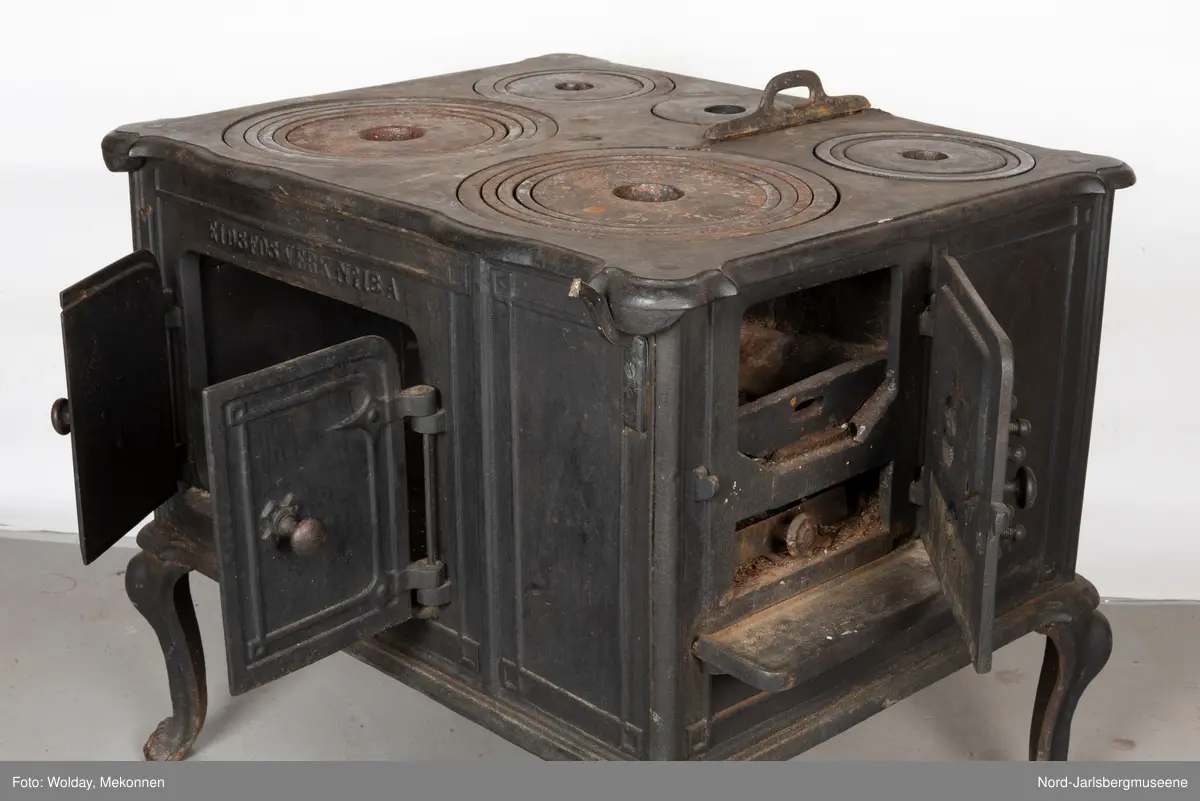 Stor, firkantet komfyr med dobbel ovnsdør til stekeovn? med støpte knotter. Enkel dekor (stilisert art nouveau). Sidedør til stekeovn. Fem kokeplater.  Bena svungne.