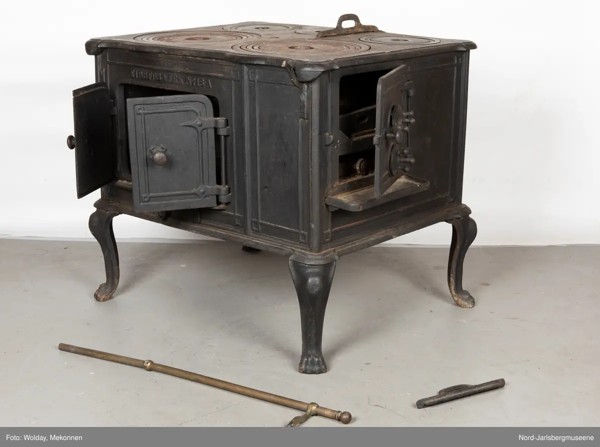 Stor, firkantet komfyr med dobbel ovnsdør til stekeovn? med støpte knotter. Enkel dekor (stilisert art nouveau). Sidedør til stekeovn. Fem kokeplater.  Bena svungne.