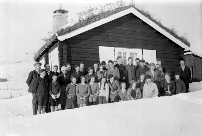 Tømmerhytte med hvite vindskier og vinduslemmer. Torvtak. Eleveklasse fra Gudbrandsdal fylkesskole foran. Vinter.