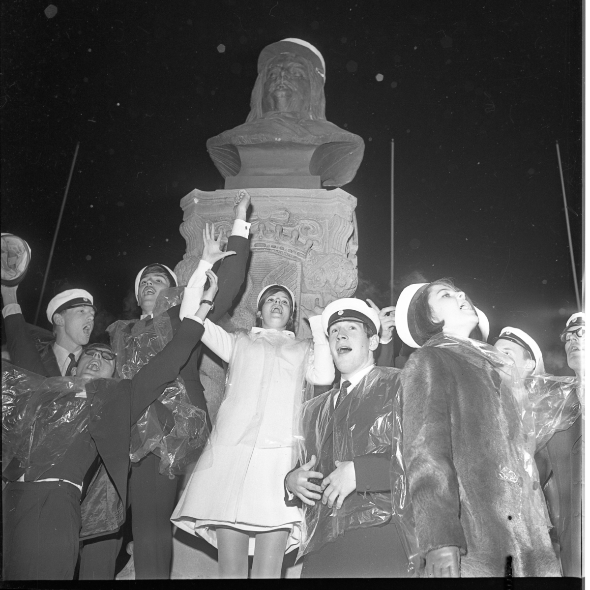 Studenter vid Grännaskolan, 1967, samlade under Brahebysten på Gränna torg. De vinkar och sjunger, troligen. De har transparent plastskydd över kläderna. Brahe har fått en studentmössa på huvudet.