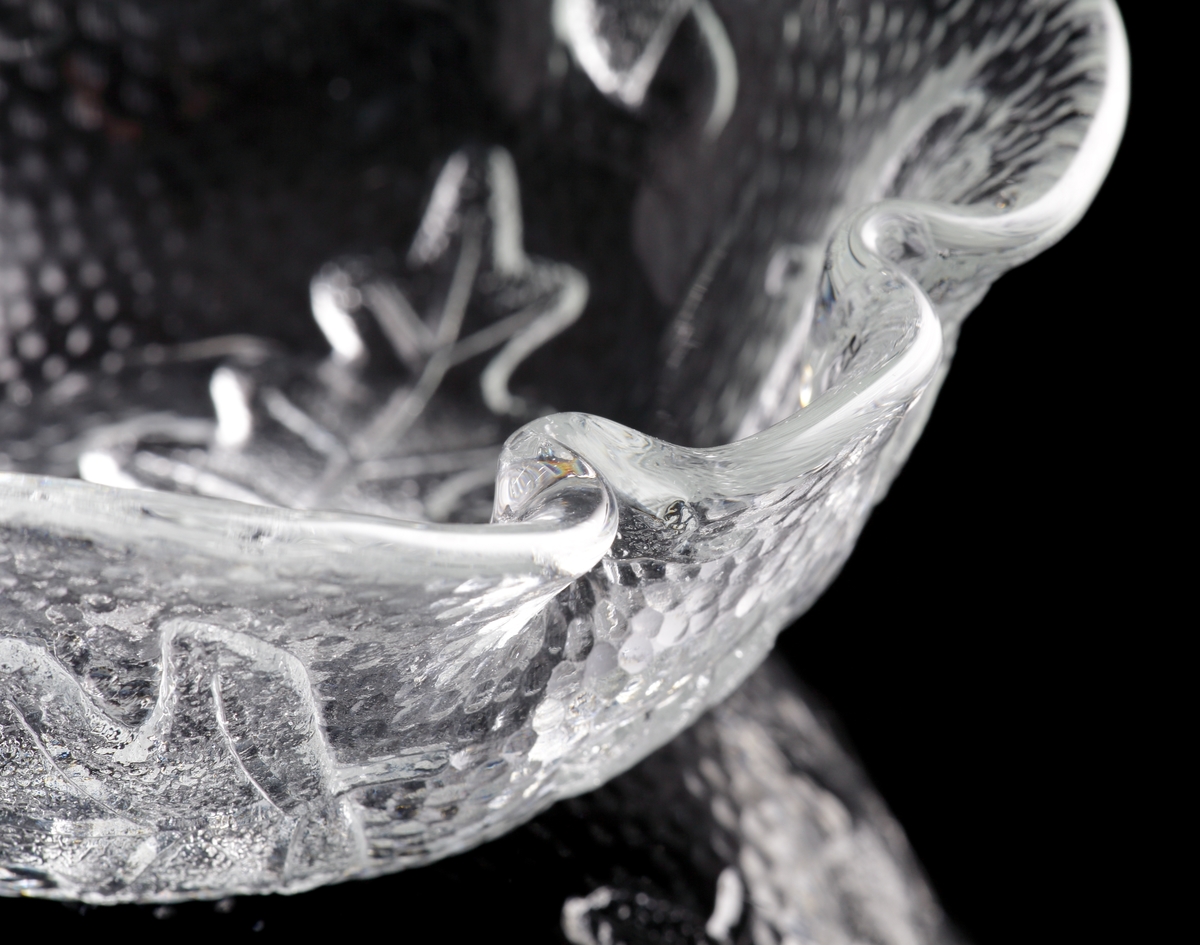 Asymmetriskt skål i ofärgat glas med reliefdekor av eklöv och ekollon.
Skålen är först gjuten till en "skiva" genom att man hällde ut glas över ett underlag med nämnda ekmönster. Sedan formades skålen i varmt tillstånd.