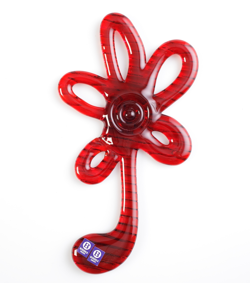 Glas som "ringlats" till ett blommotiv. Tekniken är densamma som Elving Conradsson använde till sina välkända namnskyltar.
Två st blåa fyrkantiga etiketter med en vit prick med ett "B" i mitten och under detta "BERGDALA SWEDEN".
Tillhörande förpackning i svart papp med "ofärgad" insida. På framsidan av förpackningen en plastruta varigenom glasblomman syns. Text på förpackningen: "BERGDALA GLASBRUK GRUNDAT 1889 / HANDMADE / Vacker väggdekoration / Beautiful walldecoration".