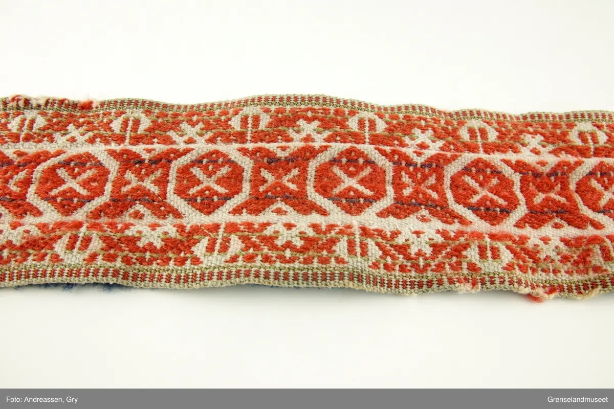 Vevd på grindvev i tre deler som er sydd sammen på langs. Sidestykkene har likt mønster. Bunn i hvitt bomullsgarn, dekor i rødt og litt blått plukk. Renningsrips. Lukning består av skinnhempe i den ene enden og skinnsnor i den andre enden. Beltet er foret med blått ullstoff.