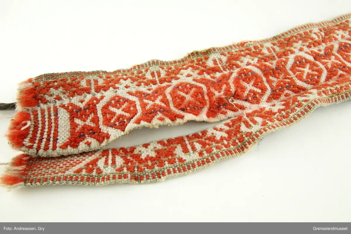 Vevd på grindvev i tre deler som er sydd sammen på langs. Sidestykkene har likt mønster. Bunn i hvitt bomullsgarn, dekor i rødt og litt blått plukk. Renningsrips. Lukning består av skinnhempe i den ene enden og skinnsnor i den andre enden. Beltet er foret med blått ullstoff.