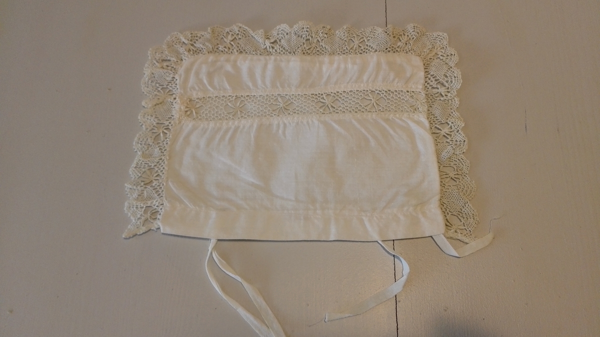 Sydd av kvitt bomullslereft med mellomverk av 2 cm brei blonde. Kanta med 3 cm brei tunga blonde kring 3 kantar. 2 sett bendelband i åpning på eine langsida.