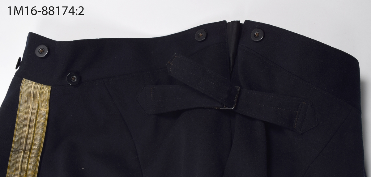 Uniformsbyxa, till lantmäteriuniform för kommisionslantmätare, av mörkblått kläde med guldrevärer längst sidorna. Sprund baktill i midjehöjd, med hank för upphängning av byxorna. Nio mörkblå knappar längs midjan. Gylften knäpps  med sex mörkblå knappar samt en hake och hyska. Fodrade upptill med beiget sidentyg med grå ränder. En ficka på vardera sida, med fickpåsar av beige bommullssatäng. Baktill byxspänne fodrat med grovt linnetyg. Nedtill på byxbenen hälla för foten, en saknas.