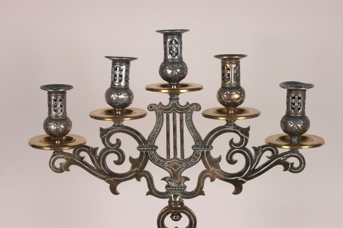 Femarmet lysestake. Rund sokkel. Fem armer springer ut fra dem midtre stolpen, som består av to lyrer. Armene og stolpen er av støpt messing. Lysholderne er i annet materiale, muligens nikkel. Dekorasjoner i hele stolpen, armene og i alle lysholderne.