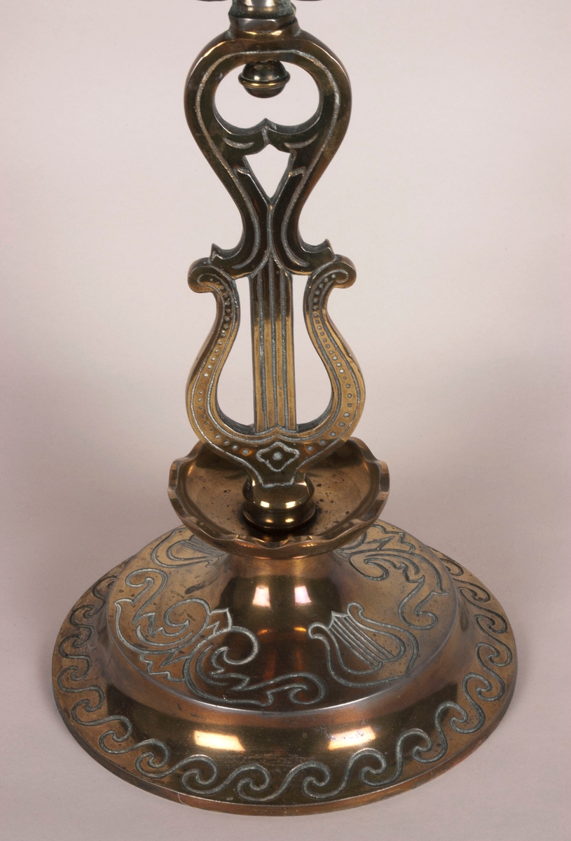 Femarmet lysestake. Rund sokkel. Fem armer springer ut fra dem midtre stolpen, som består av to lyrer. Armene og stolpen er av støpt messing. Lysholderne er i annet materiale, muligens nikkel. Dekorasjoner i hele stolpen, armene og i alle lysholderne.