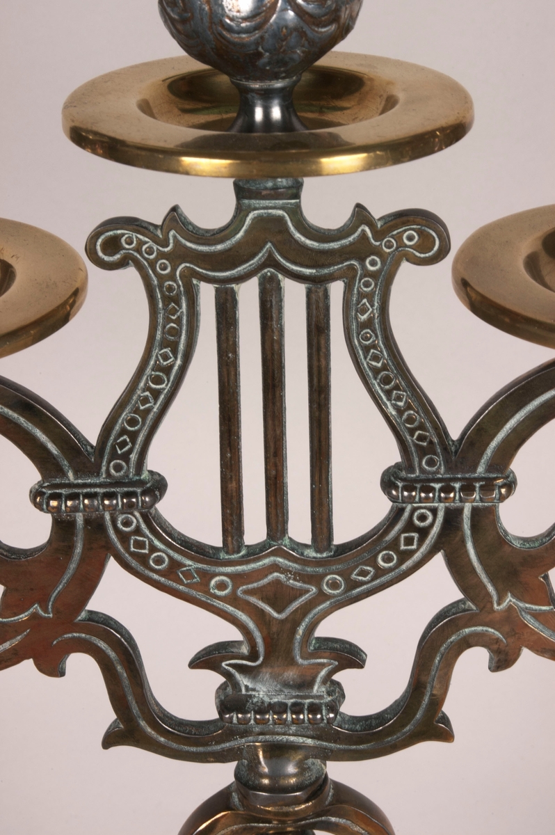 Femarmet lysestake. Rund sokkel. Fem armer springer ut fra dem midtre stolpen, som består av to lyrer. Armene og stolpen er av støpt messing. Lysholderne er i annet materiale, muligens nikkel. Dekorasjoner i hele stolpen, armene og i alle lysholderne.