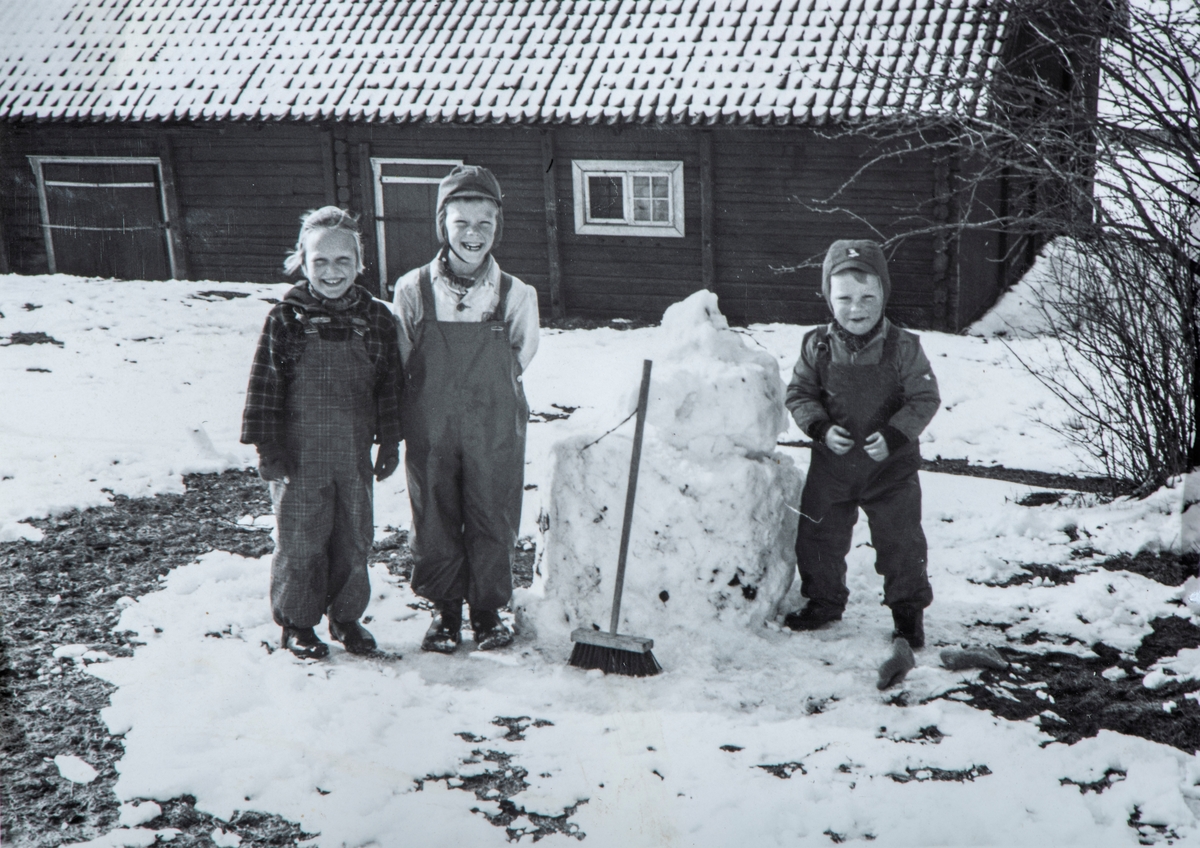 F.v. Berit Røhne, Jon Gjestvang, Erik Gjestvang, barn, barnelek, vinter, bygging av snømann. Låve, det gamle spritlageret.