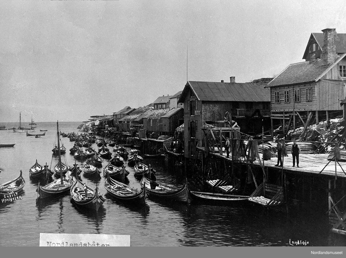 Ballstad, kaianlegg med tilhørende bygninger t.h. Flere personer på kaia. På havna ligger flere nordlandsbåter av forskjellig størrelse.