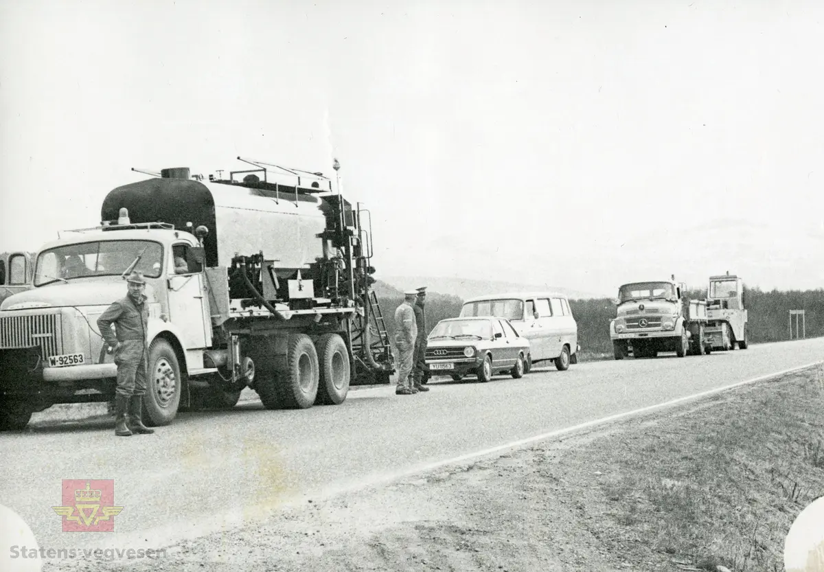 Opplæring med sprøytetank på Volvo lastebil med kjennemerke W-92563, Statens vegvesen Nordland 510-05. Testkjøring på fv på Fauskemyra 1977. En Ford Transit står bak en 1976 modell Audi 80 personbil. Mercedes-Benz lastebil foran veivalsen. Mannskapet i skyggelue og arbeidstøy (overaller) står ved bilene. 

Bilde 2) I arbeid på vegen. Mannen på sprøytebilen er godt tildekket. NVM 18-F-00414-1