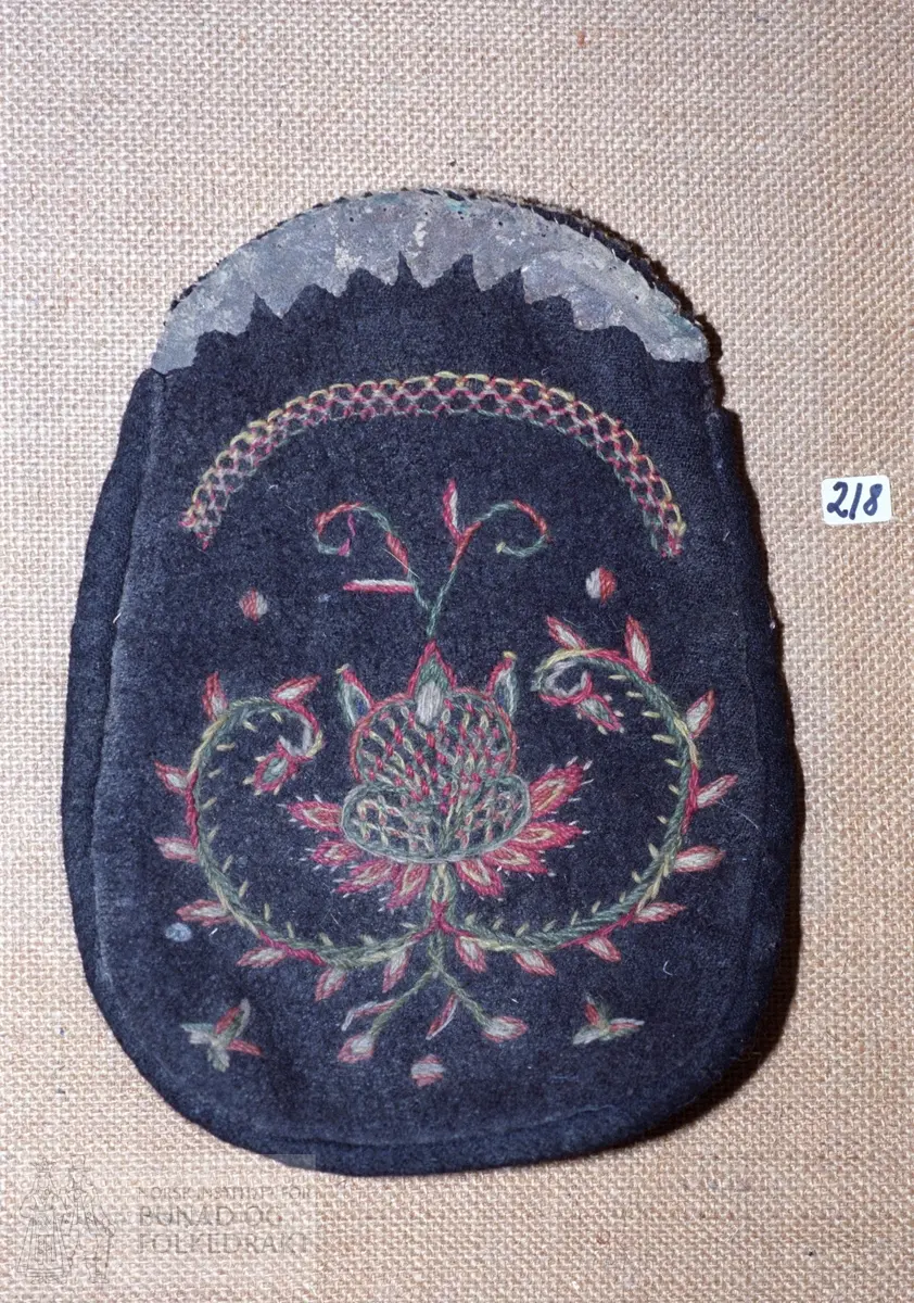 Mørkebrun vadmål på framsida pryda med rosemotiv og sauma med tynt, totråds ullgarn i raudt, grønt, gult og grått. Kontursting. "Taggekant" i skinn øvst. Skinn også på baksida. Fôra med grovt linlerret. Sauma med ubleika lingarn. Høyde:  25 cm.
