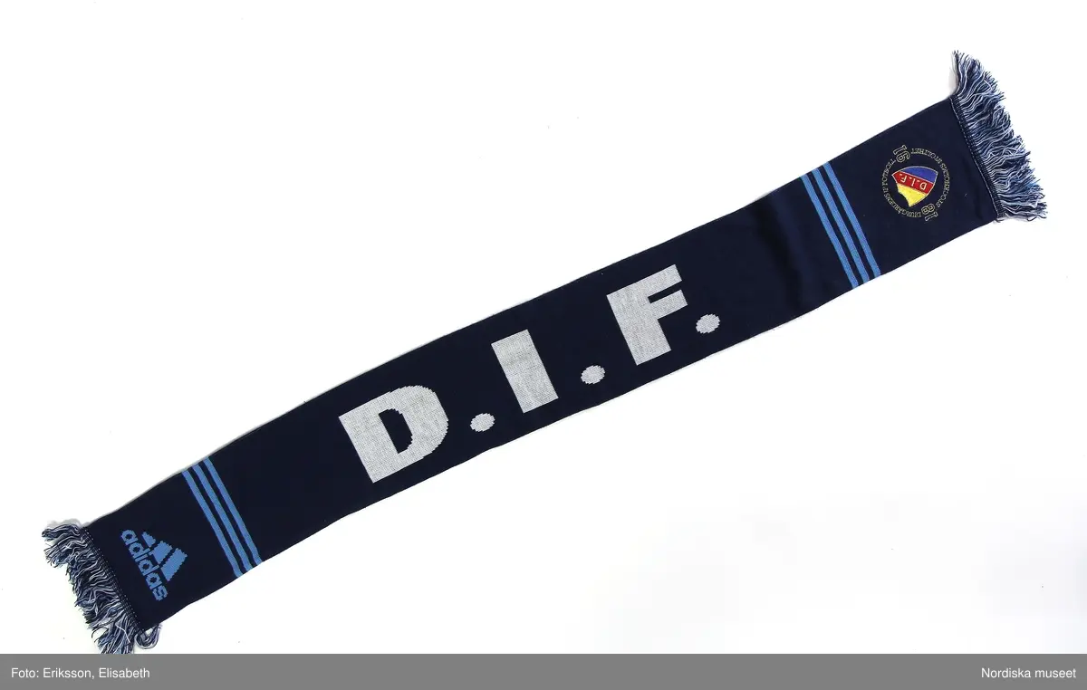 Föreningshalsduk från Djurgårdens idrottsförenings (DIF) med färgerna ljus- och mörkblått. Med fransar.
Några med Djurgårdens emblem i gult-rött-blått.
Längden mellan ca 120-160 cm.
Bredd ca 15-20 cm.

2019-02-21 Cecilia Hammarlund-Larsson/Lena Kättström Höök