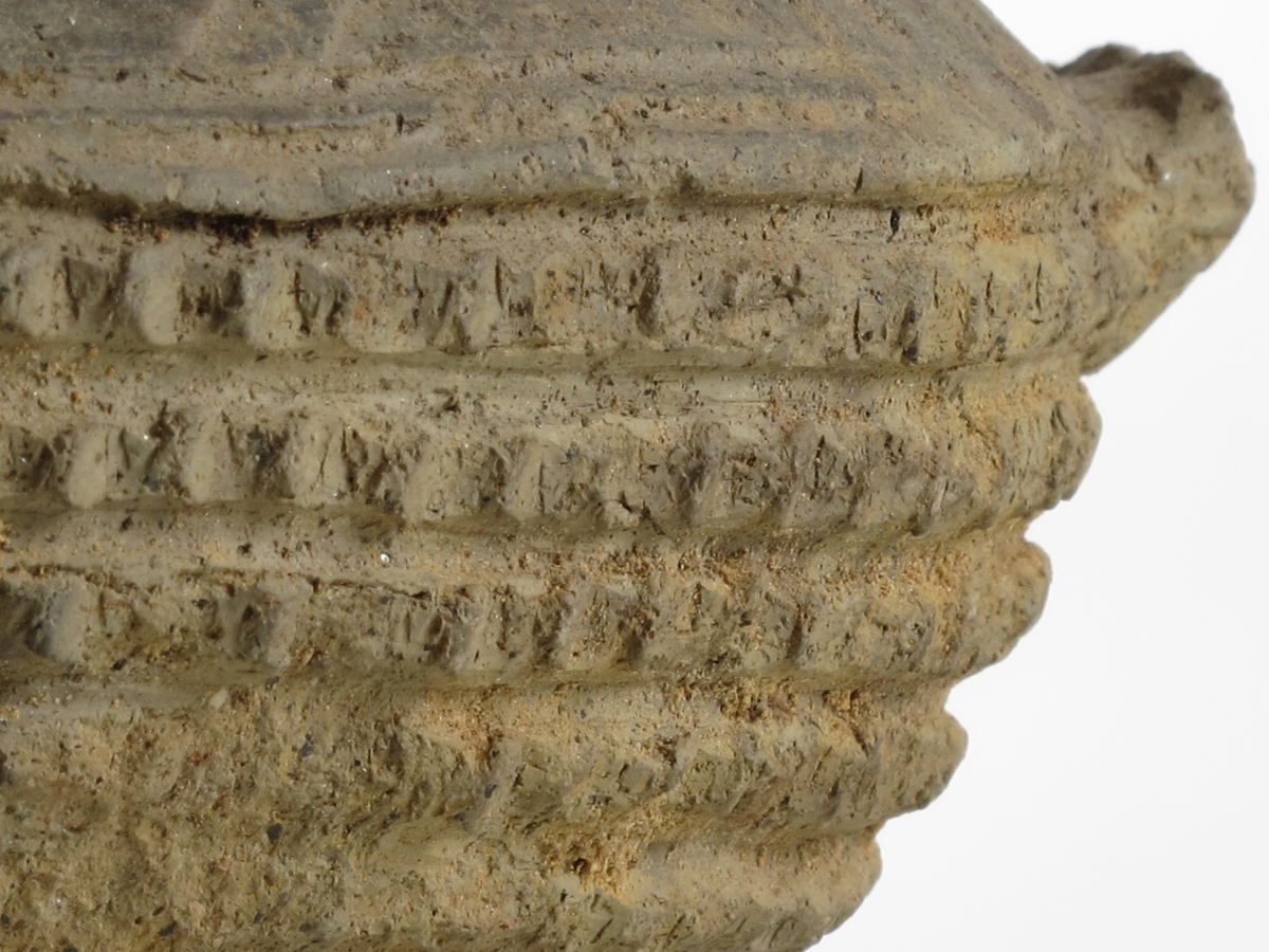 Vorte-kar med hank av lyst grålig, temmelig tynt gods av form nærmest som Bøe: anf. arb., fig. 136. Hals- og skulderornering består øverst og nederst av tre dobbelte linjebunter; over disse en punktert linje, mellom dem en hulkile og endelig parvis skråstilte linjer. Orneringen bøyer ned på begge sider av hanken. På øvre del av buken 5 rekker med vorter av samme type som anf. arb. fig. 135, men mer tettstilte. Nedre del uornert. Bunnen, som er oppdrevet, er avgrenset med en dobbeltlinje. Noen skår mangler ved randen,hanken er avslått og overflaten noe avskallet, ellers bra bevart.