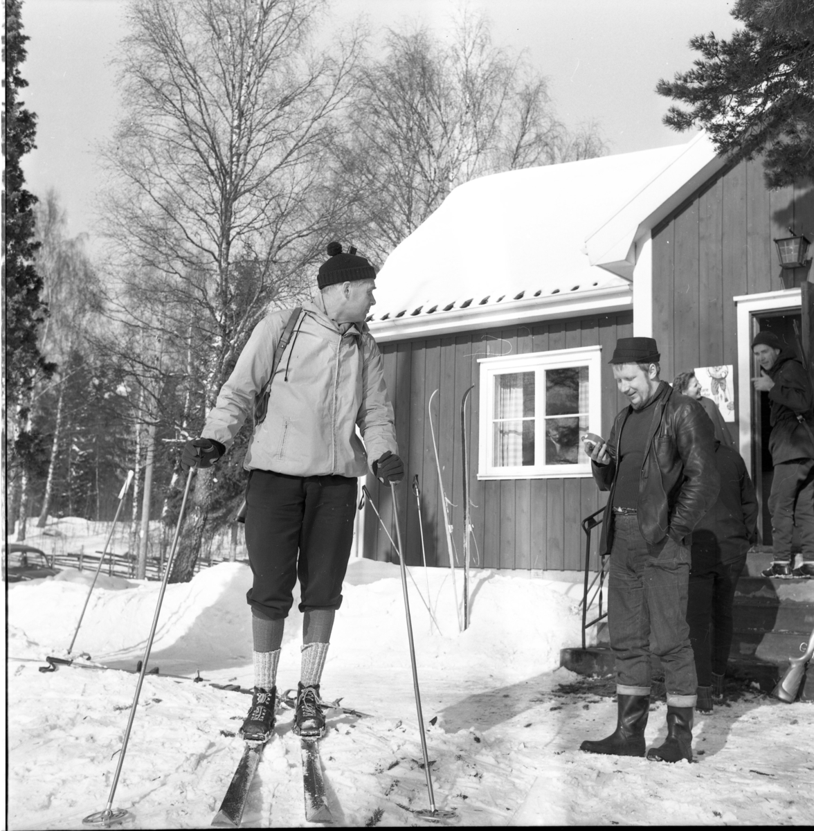 Skidskytte i Örserum. Den skidande skytten är Bengt Lycken. Han talar med en man till höger som är funktionären Anders Palmér, möjligen med ett tidtagarur i handen. De står utanför Örserums hembygdsgård. Båda är aktiva i Örserums Skytteförening. Skidor står nedstuckna i snön utanför stugan och ett gevär är lutat mot farstun.Banan sträckte sig från Hembygdsgården till skjutbanan.
