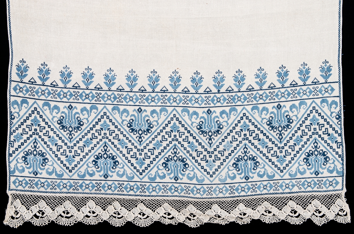 Serveringsbordsduk i vitt linne, broderad med blått korssömsbroderi i två nyanser. Framkanten i knypplad spets.