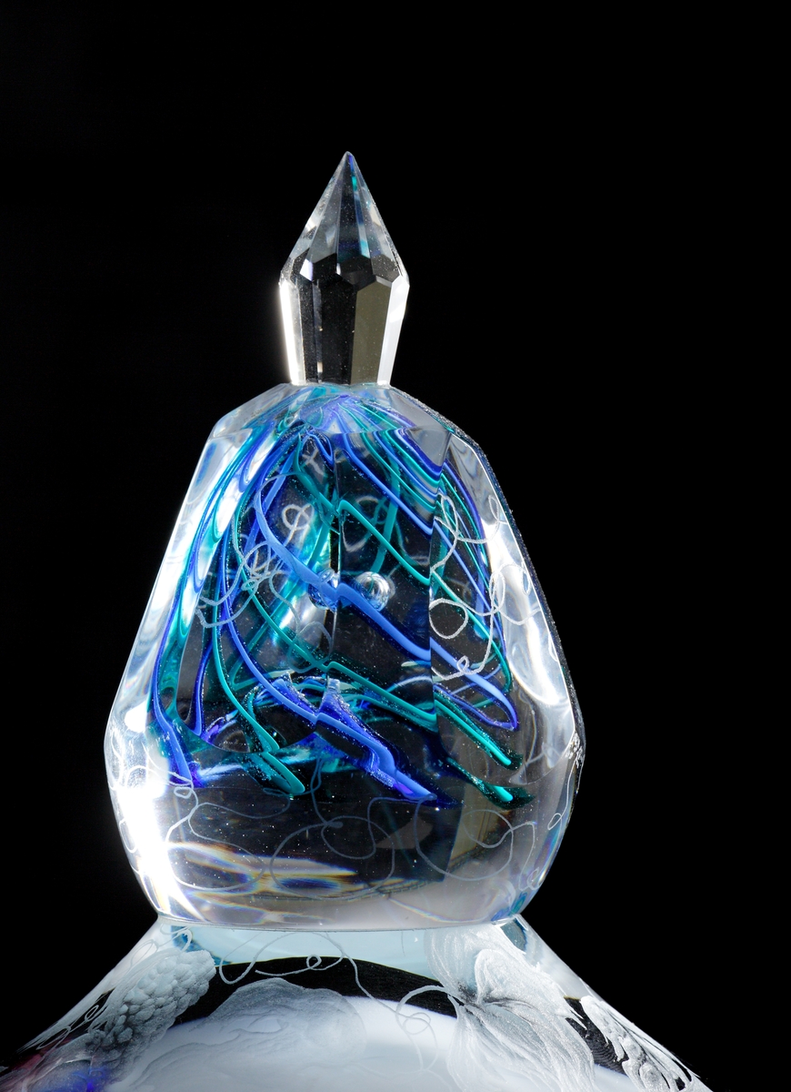 Fyrkantig glassockel. Droppformad skulptur som avslutas med en slipad knopp. I knoppen gröna och blå linjer. Skulpturen graverad med detaljerat motiv från Edens lustgård.