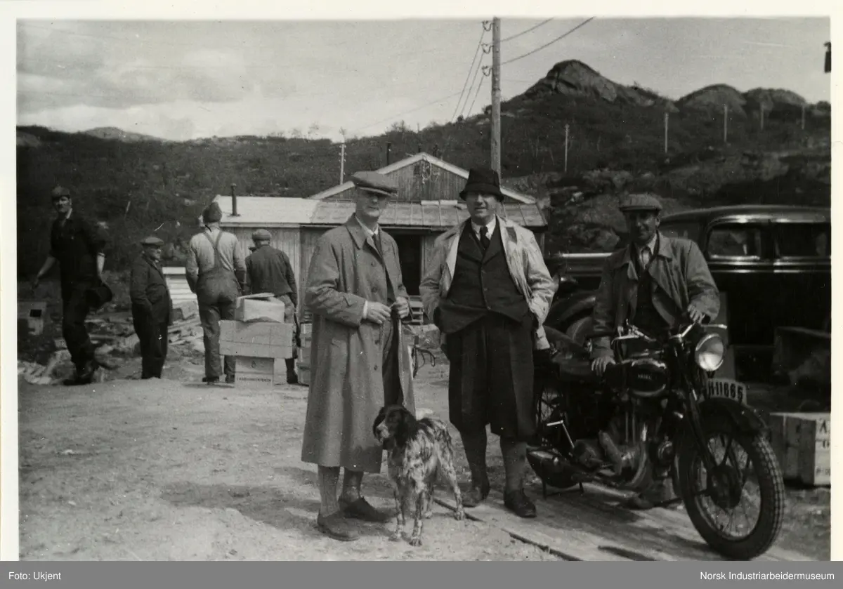 Fagerberg sag. Fagerberg sag. Mann med sikspens og ytterjakke med hund står ved siden av en mann med nikkers, dress og bøttehatt. Ved siden står en mann som holder en motorsykkel. Bak sees en gruppe arbeidsmenn