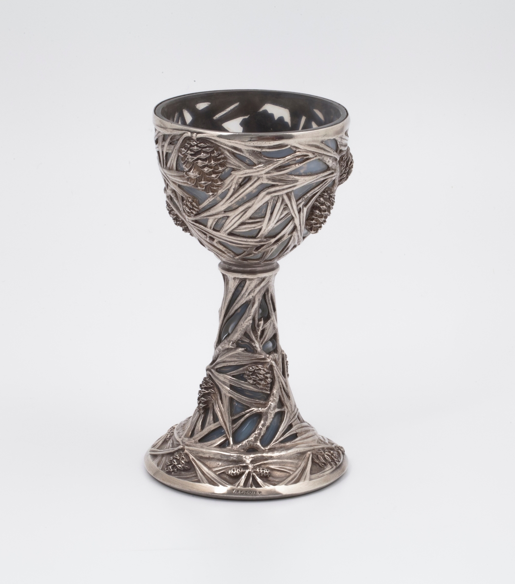 Pokal utført i støpt sølv og halvgjennomskinnelig melkehvitt opalinglass. Det gjennombrutte sølvet danner et flettverk av pinjekongler, grener og nåler i relieff, som brer seg fra den halvkuleformede kupaen og nedover den koniske stetten. Overgangen mellom disse to delene er markert med en ringformet vulst. Inne i pokalen er det innblåst opalinglass, som delvis trer frem som glassblærer på dens ytterside. På foten er det rektangulære felter med stiliserte kongler og grener i lavt relieff.