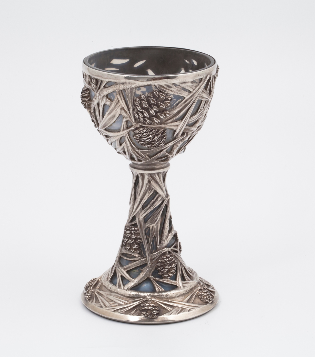Pokal utført i støpt sølv og halvgjennomskinnelig melkehvitt opalinglass. Det gjennombrutte sølvet danner et flettverk av pinjekongler, grener og nåler i relieff, som brer seg fra den halvkuleformede kupaen og nedover den koniske stetten. Overgangen mellom disse to delene er markert med en ringformet vulst. Inne i pokalen er det innblåst opalinglass, som delvis trer frem som glassblærer på dens ytterside. På foten er det rektangulære felter med stiliserte kongler og grener i lavt relieff.