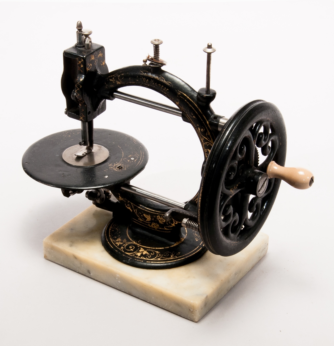 Handdriven symaskin på marmorplatta."The little Wanzer", för dubbeltrådig söm, med fram- och återgående skyttel enligt Howes patent. Märkt: R.M. Wanzer & Co. Patentees & Manufs Hamilton Canada West: Time utilizer. Trade (timglas) mark. Patented in America Gt Britain and the continent May 16 & June 17 1867".
Tillbehör: låda av trä med skjutlock och handtag, trälådans mått: 360x310x185,  Vikt:3,3kg, starkt sliten,  skyttel, 5 st spolar (2 saknas), 4 st fållvikare (saknas), 2 st ramar (?) för brodering(?), 2 st hållare (?) för brodering (?), 2 st kulor med filt (smörjkoppar ?).