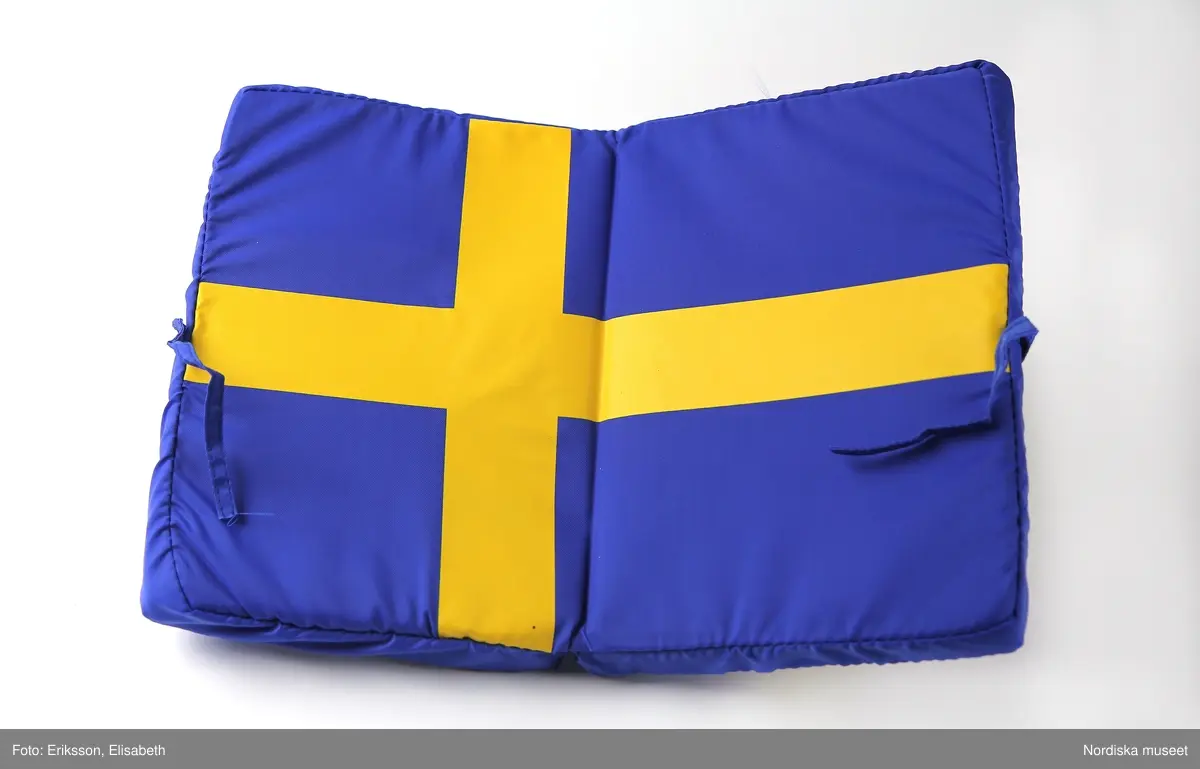 Sittdyna med svenska flaggan som dekor.  Av blått syntetmaterial med fyllning av skumgummi. På framsidan står SVERIGE i gult tryck, därovanför en version av Sveriges landslagsmärke för fotboll med texten Sverige På baksidan en svensk flagga i tryck. Dynan är uppfällbar, själva sittdynan har också svenska flaggan som dekor. Dynan kan knytas till i hopfällt skick. Oanvänd.

/ C Hammarlund-Larsson 2019-01-25