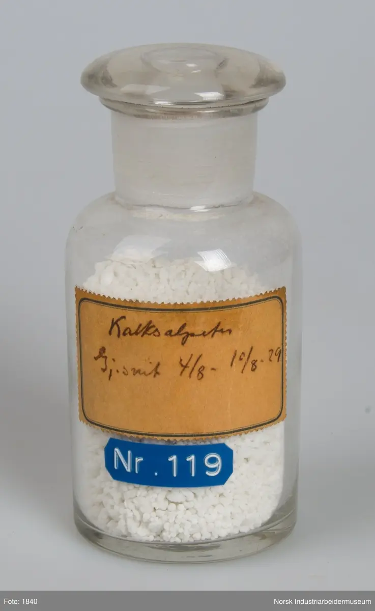 Laboratorieglas med prøve af den første produktion af kalksalpeter.
Cylindrisk laboratorieglas med slebet glasprop. Indeholder salpeter i form af små hvide perler og gryn.