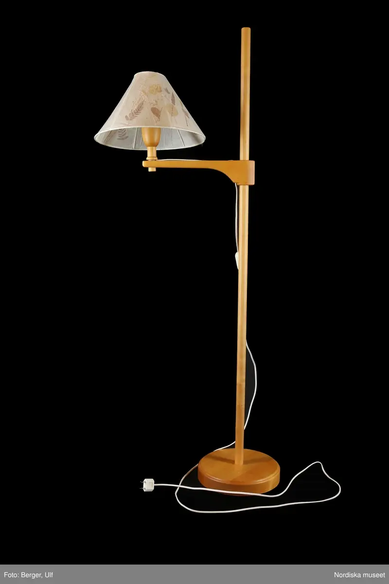 a) Golvlampa av trä, elektrisk, med höj- och sänkbar arm. Rund bottenplatta med rak, 130 cm lång vertikal stång. På stången  höj- och sänkbar arm med glödlampsfäste längst ut. b) Konisk skärm av papper med torkade pressade växter omsluter med klofattning c) matt glödlampa.
/Maria Maxén 2017-06-26