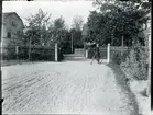 Kung Karl sn, Kungsör.
Pojke (ung man) står vid cykel på en grusplan, med staket  och villor i bakgrunden. 1913.
Text under bilden 