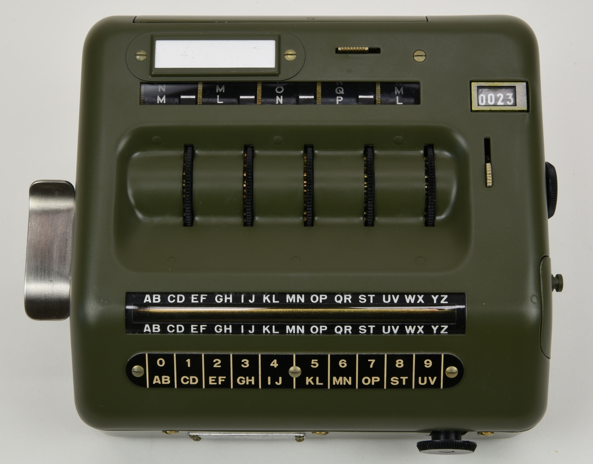 Krypteringsapparat (:1) med hölje av grönmålat aluminium i rundade former. Översidans framkant är lätt fasad och har ett avlångt fönster med alfabetet i över- och nederkant. Fönstret är till för att visa den alfabetsrulle som gav lösningen på kryptot. Under fönstret finns en svart skylt med vit text där siffrors bokstavskoder visas. På ovansidans mitt finns fem smala chifferhjul av svart bakelit i en försänkning. Hjulen är till för att ställa in chiffrets bokstäver som syns i ett fönster längst upp på apparatens ovansida. Till höger om chifferfönstret finns ett räkneverk med fyra siffror.

På apparatens framkant finns en skylt med M-nummer, serienummer och liknande och till höger ett vred av svart bakelit för att mata fram alfabetsrullen. På apparatens vänstra sida finns ett frammatningsvred av metall samt tillverkarens logotyp. På den högra sidan finns en lucka för alfabetsrullen och ett vred av bakelit för att ställa in chifferhjulaxeln. Baksidan har en större lucka för hålkortsplatsen och undersidan har små gummifötter samt en dekal med information om garantitid.

Till apparaten hör också en transportväska (:2) av formgjuten och genomfärgad gummi. Väskan är grön och har en vävd bärrem fäst med mässingsskruv. Locket är fäst med ett avlångt, nitat gångjärn och har ett låsspänne i mässing. På locket finns tre kronor och apparatens M-nummer i relief och på väskans undersida finns väskans M-nummer. Insidan av väskan är formad efter kryptoapparaten för att ge så mycket skydd som möjligt.

M-nummer: M 3858-301010
Tillverkningsnummer: 22749

Väskans M-nummer: M 7038-045010