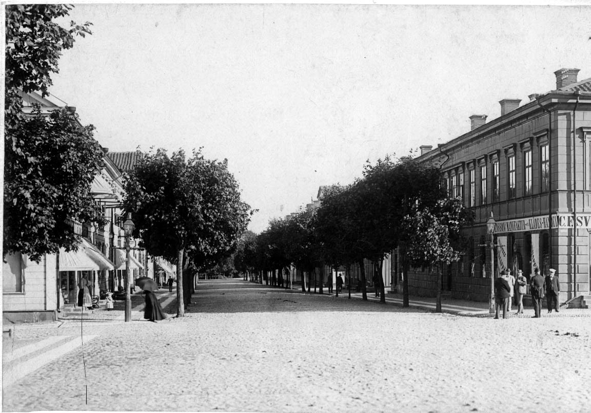Stadsbild från Eksjö. Södra storgatan kantad med lövträd och bebyggelse. En kvinna i mörka kläder med parasoll hastar till vänster. Mittemot står ett antal män utanför C E Svenssons Manufaktur och Klädeshandel.