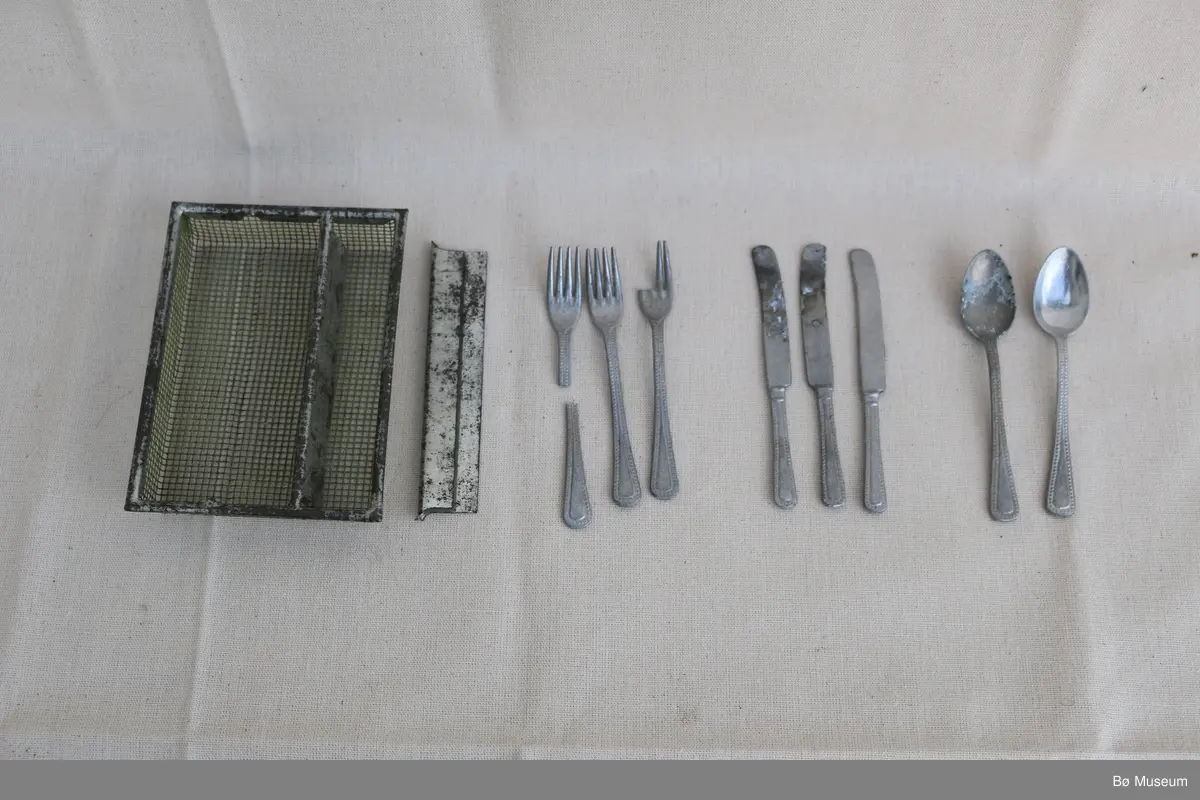 Lite leikebestikk for born, med lita bestikkorg. I bestikkorga var det opphaveleg tri kammer, men det eine skiljet er brote av og ligg laust i det eine rommet.  Ramma og skilja til bestikkorga er opphaveleg krombelagt/kromlakkert metall, og nettingen, altså botnen og ytterveggane, er grønmalt netting i metall. Bestikket er av aluminium. To av gafla er øydelagde.

a - Spiseskjeier, 2 stk.
b - Knivar, 3 stk.
c - Galflar, 3 stk. Ein gaffel er heil, ein gaffel manglar to tenner, og ein gaffel er brote i to på midten.
d - Bestikkorg