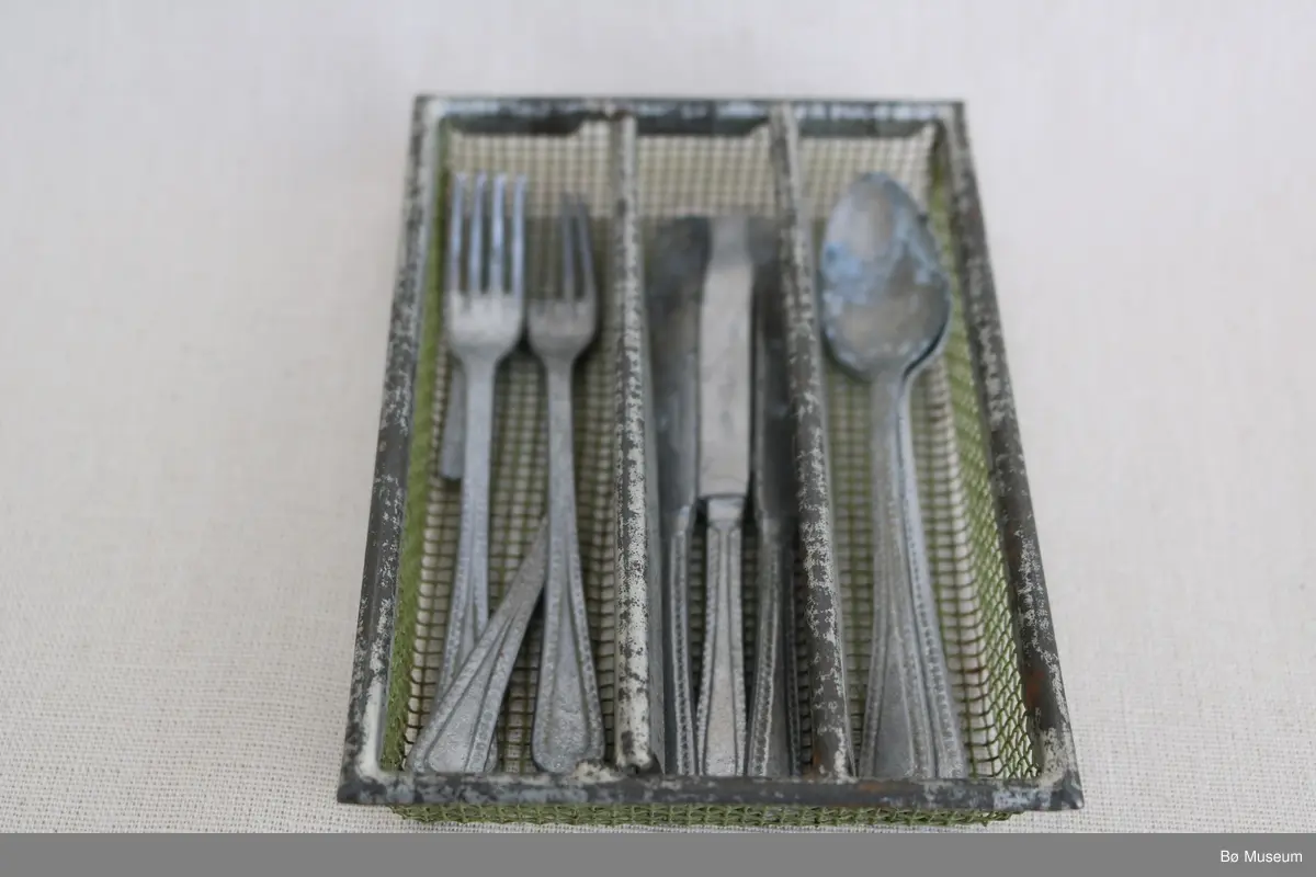 Lite leikebestikk for born, med lita bestikkorg. I bestikkorga var det opphaveleg tri kammer, men det eine skiljet er brote av og ligg laust i det eine rommet.  Ramma og skilja til bestikkorga er opphaveleg krombelagt/kromlakkert metall, og nettingen, altså botnen og ytterveggane, er grønmalt netting i metall. Bestikket er av aluminium. To av gafla er øydelagde.

a - Spiseskjeier, 2 stk.
b - Knivar, 3 stk.
c - Galflar, 3 stk. Ein gaffel er heil, ein gaffel manglar to tenner, og ein gaffel er brote i to på midten.
d - Bestikkorg