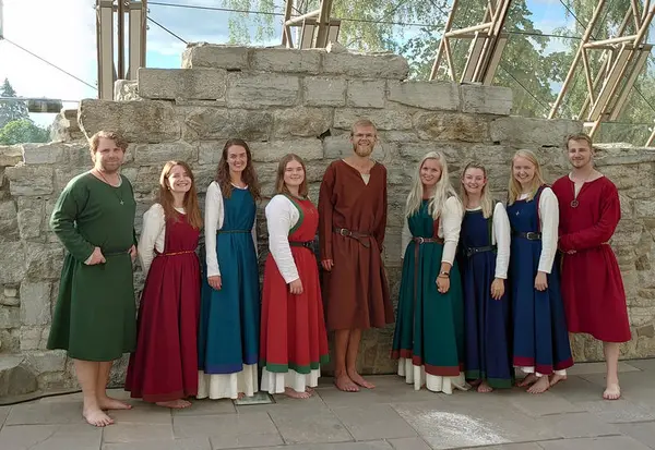 Åtte ungdommer i middelalderdrakt står pent på rekke foran ruinen av en kirkemur.