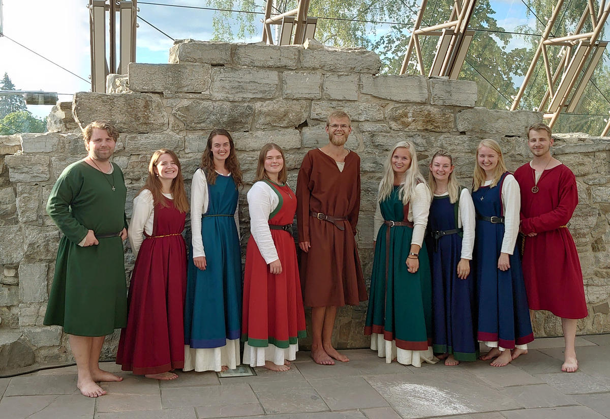 Åtte ungdommer i middelalderdrakt står pent på rekke foran ruinen av en kirkemur.