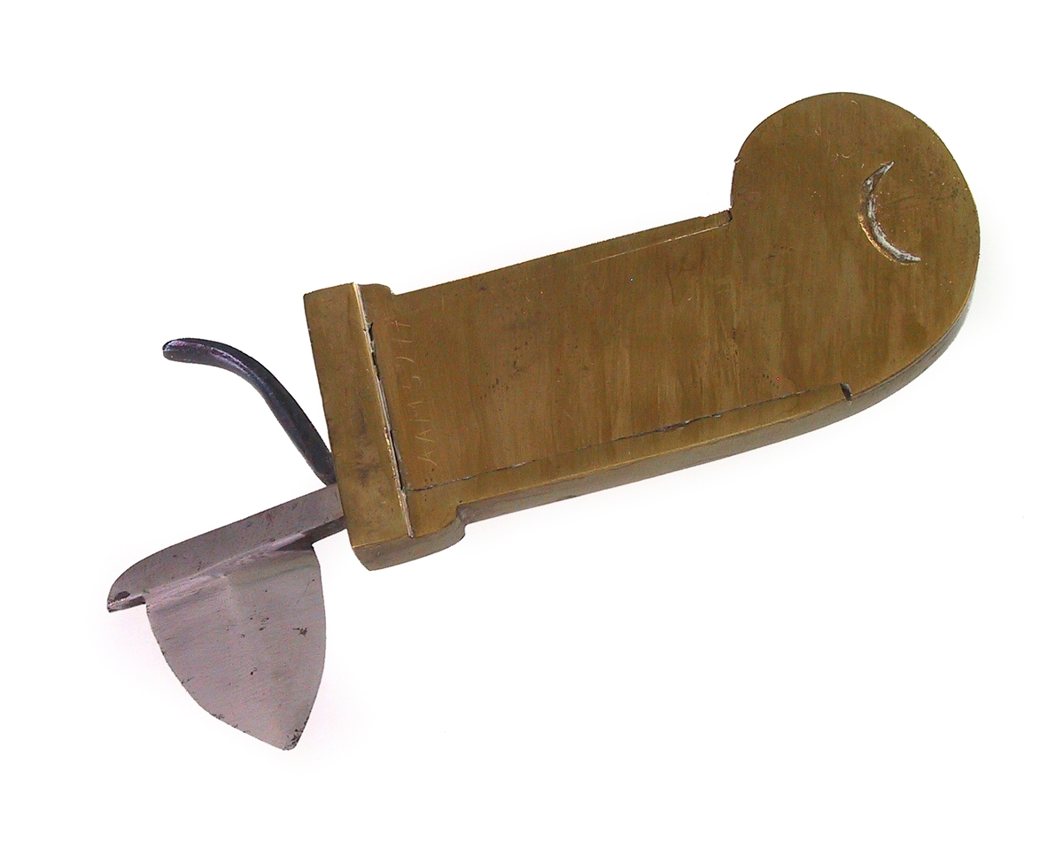 årelatingsinstrument. Messing og stål. Massiv messingplate, bred J-form. På den ene side en hengslet, svunget og tunget "avtrekker" med fjær som gjør at den kan løftes opp og  ned og med en tapp i den ende som hviler mot platen. Øverst en fjær og et skarpt tveegget knivblad av form som en halv ellipse. På baks. nederst en halvsirkelformet nedsenket bue. Et trykk på avtrekkeren, med fjæren spent, får kniven til å hugge.