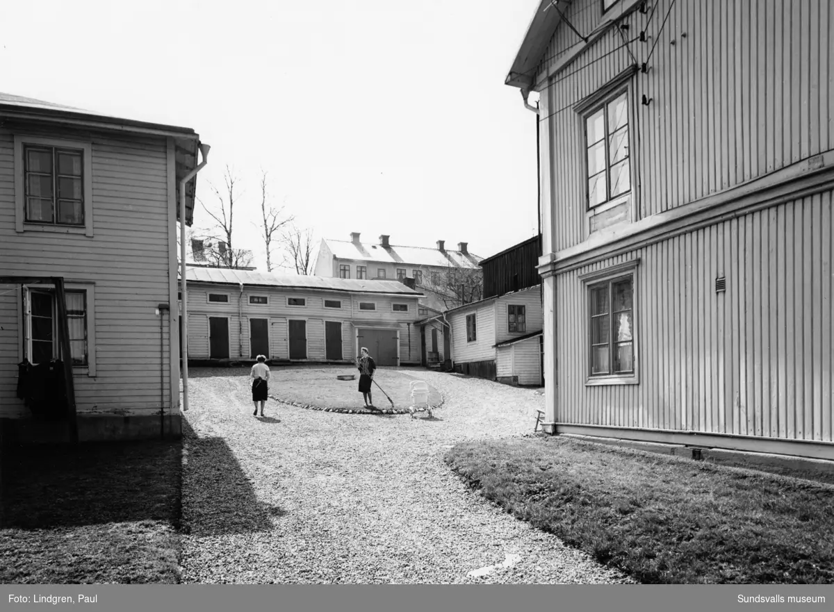 Nygatan 19. Fastigheten uppfördes 1895 av handlaren M. A. Fürst som även där bedrev handelsrörelse (speceriaffär) i det tillhörande huset. Byggnaderna är uppförda av timmer som spönats.1901 sålde Fürst fastigheten till torparen Erik August Sjölund som innehade densamma ett par år varefter Fürst åter fövärvade egendommen. 1907 övertogs fastigheten av inspektoren på Rosenborg A. J. Berg. Rörelsen drevs fram till första världkriget av fru Johanna Berg. I gårdshuset inrättades en elektrisk mangel, som efter båda makarnas frånfälle övertogs av en dotter och som alltjämt var i full verksamhet. t.o. m. 1960 då även denna nedlades. På denna bild ses Gertrud Eriksson och Helga Lindgren på gården.