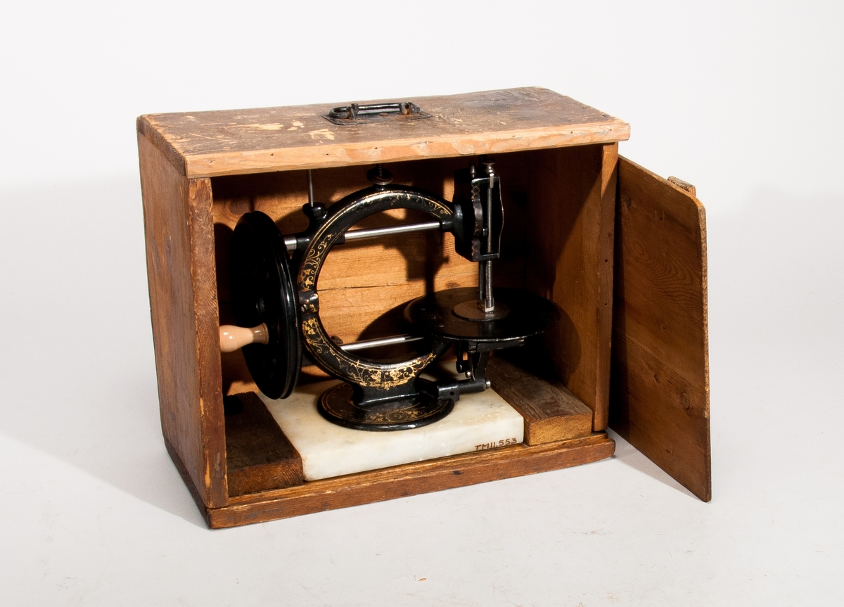 Handdriven symaskin på marmorplatta."The little Wanzer", för dubbeltrådig söm, med fram- och återgående skyttel enligt Howes patent. Märkt: R.M. Wanzer & Co. Patentees & Manufs Hamilton Canada West: Time utilizer. Trade (timglas) mark. Patented in America Gt Britain and the continent May 16 & June 17 1867".
Tillbehör: låda av trä med skjutlock och handtag, trälådans mått: 360x310x185,  Vikt:3,3kg, starkt sliten,  skyttel, 5 st spolar (2 saknas), 4 st fållvikare (saknas), 2 st ramar (?) för brodering(?), 2 st hållare (?) för brodering (?), 2 st kulor med filt (smörjkoppar ?).