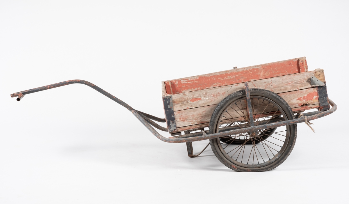 Tralle på to gummihjul. Rektangulær kasse dannet av liggende plank, en manglende kant på kortende. Metallforsterkning på alle hjørner. Understell dannet av  bøyde jernrør, hvor ett danner håndtaket. Tverrliggende jernrør til håndtering.