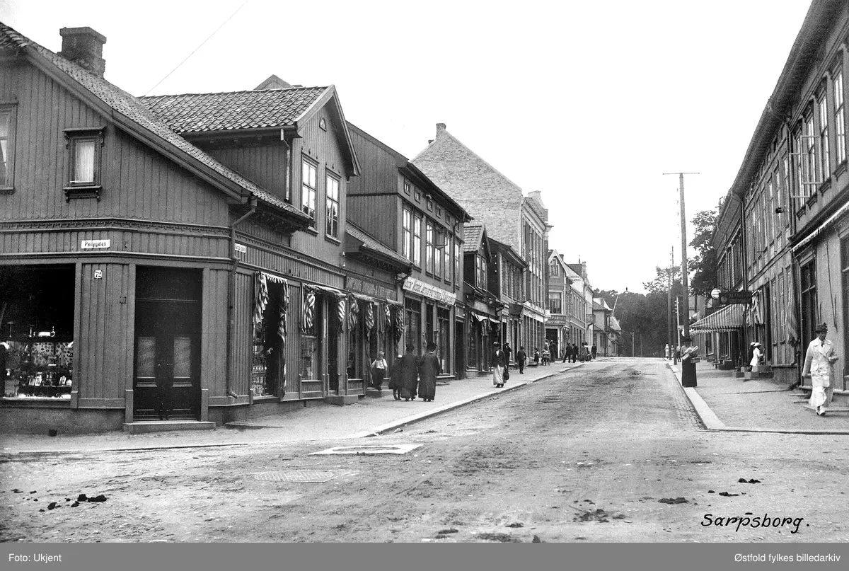 Glengsgata i Sarpsborg i krysset med Pellygata i forgrunnen, ca. 1915. Postkort. Til venstre Sarpsborg Symaskin & Cykelforretning, innehaver Th. Næss, salg av Gritsner-sykler og symaskiner, forretninga oppretta i 1914. Vegg i vegg på høyre side ligger Oscar Mostad Jernvareforretning. 
Hjørnegården til venstre i bakgrunnen er St. Marie gate 98 med Sarpsborg Broderi- og Modeforretnig, innehaver Guri Iversen. Urmaker til høyre: M. Opsahl.